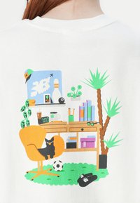 T-shirt blanc avec un graphique coloré d'un espace de travail créatif, présentant une chaise orange, un chat noir, des plantes en pot et une horloge.