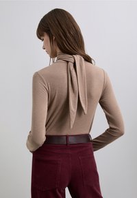 Bluză maro cu mâneci lungi și croială mulată, cu detaliu de nod la gât, asortată cu pantaloni maro închis din catifea cu buzunare la spate și curea maro.