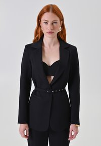 Blazer nero su misura con scollatura a V profonda, vita aderente e cintura decorata con hardware argentato. Orlo inferiore leggermente svasato.