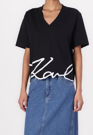 Kobieta ubrana w czarny T-shirt z dekoltem w serek z białym napisem i wysokim stanem niebieskiej spódnicy dżinsowej, stojąca na jednolitym tle.