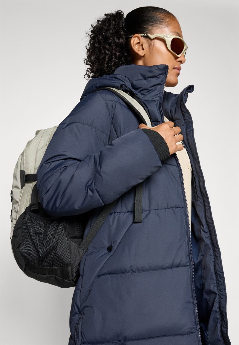 Femme aux cheveux bouclés en queue de cheval, portant des lunettes de soleil beiges, un manteau rembourré bleu marine, et portant un sac à dos gris clair et noir.