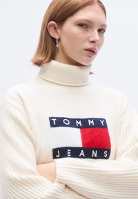 Pull à col roulé côtelé blanc avec un grand patch logo portant l'inscription "TOMMY JEANS" en couleurs bleu marine, rouge et blanc sur le devant.