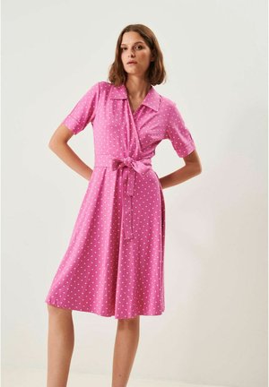 Kvinde iført en knælang pink kjole med hvide polkaprikker, korte ærmer, krave og et bundet bælte i taljen, stående foran en ensfarvet baggrund.