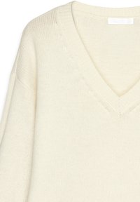 V-neck knitted sweater in cream, featuring a textured stitch pattern and ribbed neckline. The sleeves are wide and slightly oversized.
