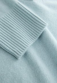 Gaiši zils adīts džemperis ar ribotu piedurkņu dizainu, mīkstu tekstūru un gludu audumu ar smalku spīdumu.