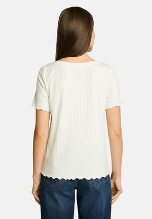 Femme aux cheveux bruns lisses portant un haut blanc texturé à manches courtes et un jean bleu en denim, vue de dos sur fond blanc.
