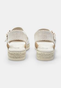 Hvide flettede sandaler med en struktureret rem, åben hæl og espadrille-stil sål. Har en gyldent spænde på ankelremmen.