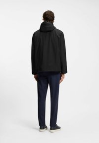 Black waterproof jacket with a hood, designed with a relaxed fit. Paired with navy trousers and dark shoes. Smooth texture, minimal detailing.