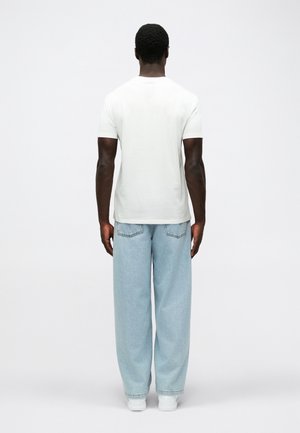 Mann, der mit dem Rücken zur Kamera steht, trägt ein schlichtes weißes T-Shirt, hellblaue Jeans mit weitem Bein und weiße Turnschuhe vor weißem Hintergrund.