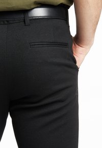 Zwarte, op maat gemaakte broek met een gladde textuur, voorzien van een achterzak en een glanzend zwarte riemdetail. Strak, minimalistisch ontwerp.