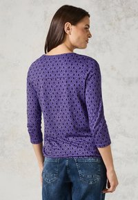 Violetti paita, jossa on timanttikuvio, 3/4-hihat ja pyöreä kaula-aukko, yhdistettynä sinisiin farkkuihin, joissa on takataskut.