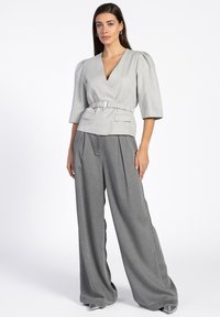 Lichtgrijze wikkelblouse met korte pofmouwen, een ceintuur in de taille en twee voorzakken, gecombineerd met brede grijze broekspijpen en zilveren hakken.