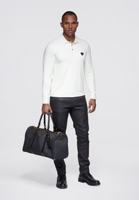 Weißes langärmeliges Polohemd mit schwarzem Emblem, schwarze Jeans und eine schwarze Reisetasche mit Muster und goldenen Beschlägen. Schwarze Stiefel.