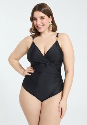 Nainen, joka käyttää mustaa yksiosaisetta uimapukua, jossa on kultaiset ketjuhihnat, hymyilee, seisoo käsi lanteilla yksiväristä taustaa vasten.