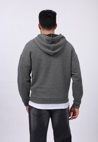 Gray waffle-textured hoodie with a hood, ribbed cuffs, and hem. Worn over a white shirt, paired with dark jeans, viewed from the back.