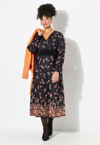 Schwarzes Blumenkleid mit langen Ärmeln, gerafften Bündchen und einem schwarzen Taillengürtel. Das Model hält eine orange Jacke, schwarze Strumpfhosen und hohe Schuhe.