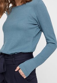 Haut à manches longues texturé de couleur bleu doux avec col rond. Le tissu présente un motif en losange subtil. Comprend des poignets ajustés.