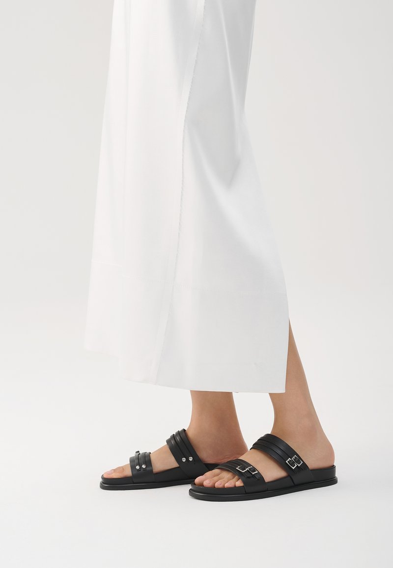 Schwarze Riemchensandalen mit zwei breiten Riemen, silbernen Akzenten und flacher Sohle. Das Design verfügt über eine glatte Oberfläche und einen offenen Zehenstil.