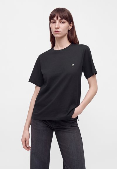 Femme aux cheveux bruns lisses portant un t-shirt noir uni à manches courtes et un jean foncé, debout avec une main dans la poche devant un fond uni.