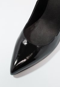 Escarpin à talon haut en cuir verni noir avec un bout pointu et une texture lisse. Le contour intérieur correspond à l'extérieur, accentuant le design élégant.