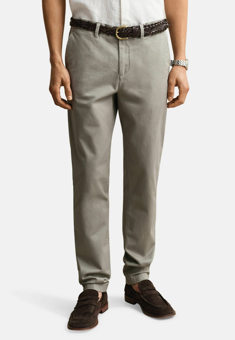 Homme portant un pantalon beige avec une ceinture tressée marron foncé, une chemise blanche, une montre argentée et des mocassins en daim marron foncé.