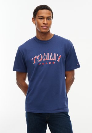 Jeune homme debout, les mains dans le dos, portant un t-shirt Tommy Jeans bleu marine et un jean foncé sur fond blanc.