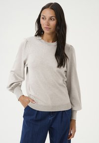 Pull léger beige avec manches longues, encolure arrondie et ourlet côtelé. Présente des manches bouffantes et une texture douce. Porté avec un jean bleu foncé.