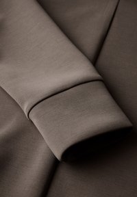 Brun tygsleeve med en slät, texturerad finish och en åtsittande mudd. Materialet har en lätt lyster och är lättviktigt.