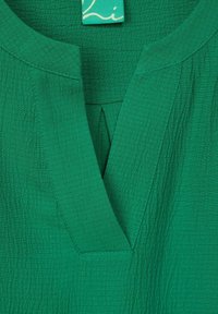 Blusa verde con texture che presenta una scollatura a V, dettagli di pieghe sottili e un'etichetta tessuta sul colletto. Tessuto liscio con un aspetto increspato.