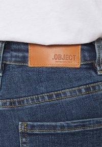 Ruskea nahkapatche, jossa on '.OBJECT keräilytuote' -labeli, sijaitsee sinisten farkkujen vyötäröllä, ja niitä on yhdistetty valkoiseen paitaan.