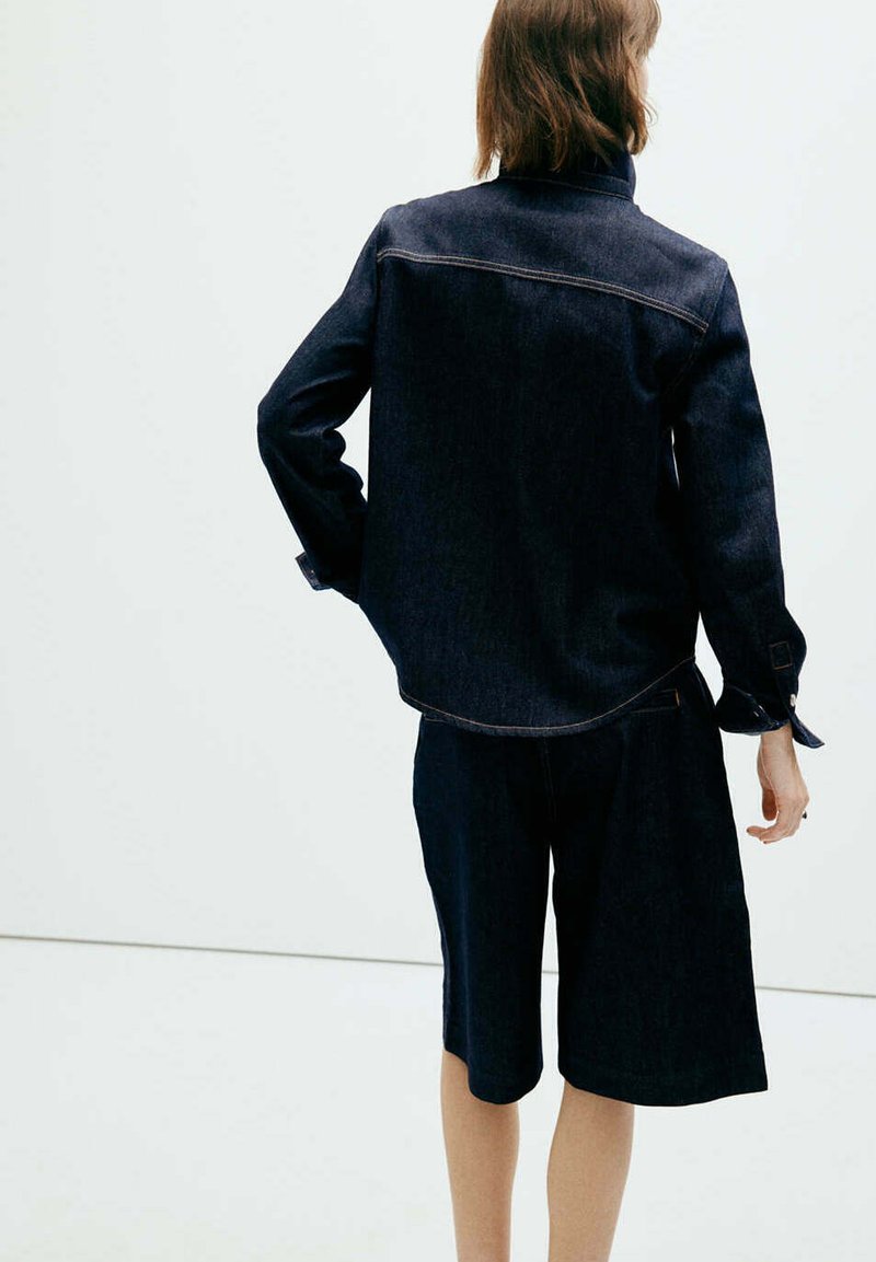 Personne aux cheveux mi-longs portant une veste en jean foncée et un short assorti jusqu'aux genoux, debout avec une main sur la hanche sur un fond uni.