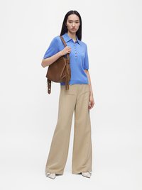 Femme portant un polo bleu à manches courtes, un pantalon beige à jambes larges, des chaussures blanches et un sac à main en cuir marron avec pompons sur l'épaule.