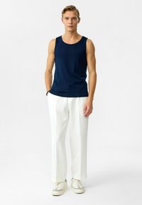 Marineblauw mouwloos shirt, witte brede broek met verticale strepen en crèmekleurige sneakers. De outfit heeft een ontspannen fit en gladde texturen.