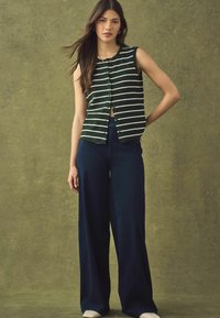 Grün-weiß gestreiftes ärmelloses Oberteil mit durchgehender Knopfleiste, kombiniert mit weit geschnittenen, navyblauen Hosen. Glatte Stoffstruktur, lässiges Design.