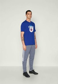 Blå kortermet t-skjorte med "FRANCE" grafikk, grå joggebukser med hvite striper, og svarte treningssko. Modellen står.