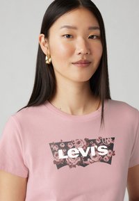 T-shirt en coton rose pâle avec un design floral et le logo "Levi's" en blanc, sur fond sombre. Manches courtes, col rond.