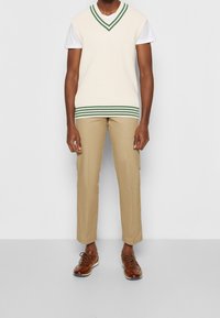 Cremefarbener, gerippter, ärmelloser Pullover mit grünen Streifen am Saum, getragen über einem weißen T-Shirt, kombiniert mit beigen Hosen und braunen Schnürschuhen.