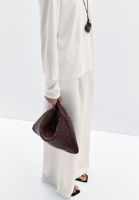 Vävd bourgogneväska med triangulär form som hålls i en hand. Outfiten har en löst sittande vit topp och matchande byxor. Platta svarta sandaler.