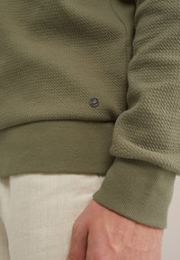 Olivno zelena majica s kapuljačom iz teksturirane pletene tkanine in rebrastimi manšeti. Ima majhen črn logotip na robu. Spodaj so vidni svetli hlače.