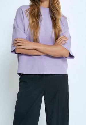 Kobieta z długimi włosami, ubrana w luźny lawendowy sweter z krótkim rękawem i czarne spodnie, z założonymi na siebie rękami na tle jednolitego tła.