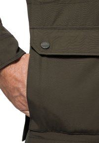 Oliwkowa kurtka z teksturowanej tkaniny. Posiada kieszeń na zamek błyskawiczny oraz akcent w postaci zatrzasku na kieszeni na piersi, podkreślając funkcjonalny design.