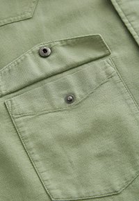 Tessuto verde con texture che presenta una tasca impreziosita da un bottone a scatto argentato, evidenziando cuciture dettagliate e un motivo leggermente in rilievo.
