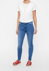 Blaue Skinny-Jeans kombiniert mit einem weißen T-Shirt und roten Rutschschuhen. Die Jeans verfügen über Fronttaschen und eine mittelhohe Taille.