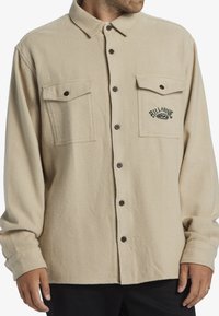 Beige fleece shirt with two chest pockets, brown buttons, and embroidered logo. Long sleeves with buttoned cuffs. Soft texture.