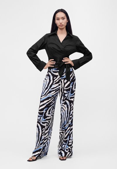 Femme debout, les mains sur les hanches, portant un chemisier cache-cœur noir et un pantalon large avec un imprimé zèbre noir, blanc et bleu, ainsi que des sandales noires.