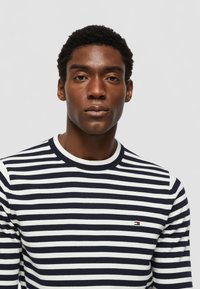 Sweater listrado em azul-marinho e branco com gola canelada, exibindo um pequeno logótipo bordado no peito. O tecido parece suave e texturizado.