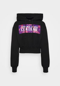 Černá zkrácená mikina s kapucí na stažení, zdobená barevným logem v růžové a fialové barvě, které zobrazuje "VERSACE JEANS COUTURE."