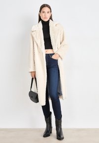 Krémový kabát z umelého kožucha s vzorom rybej kosti, prešívaný, s nadmernými klopami a jedným gombíkom. Skombinovaný s tmavými džínami a čiernymi členkovými topánkami.
