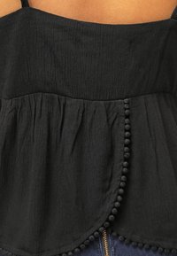 Black textured sleeveless top with pom-pom trim along the front hem, worn over blue jeans with visible gold zipper.