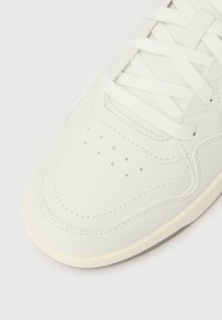 Balto sportinio batelio priekinė dalis su raišteliais, perforuotomis ventiliacijos skylutėmis ir siūtomis detalėmis ant šviesaus fono.